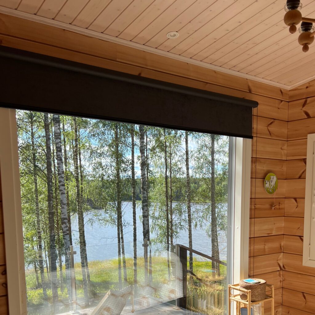 Pimentävä rullaverho, Kaihdin Sampo Pimentävä rullaverho kesämökin ikkunassa, taustalla kaunis kesäinen järvimaisema Kaihdin Sampo
